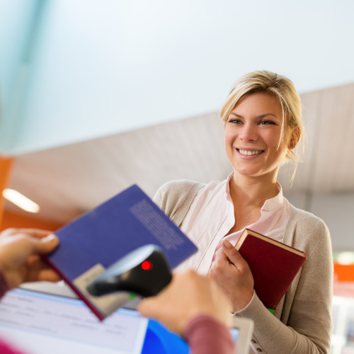 "Portrait of young female college student returning book to library, with librarian scanning barcode"